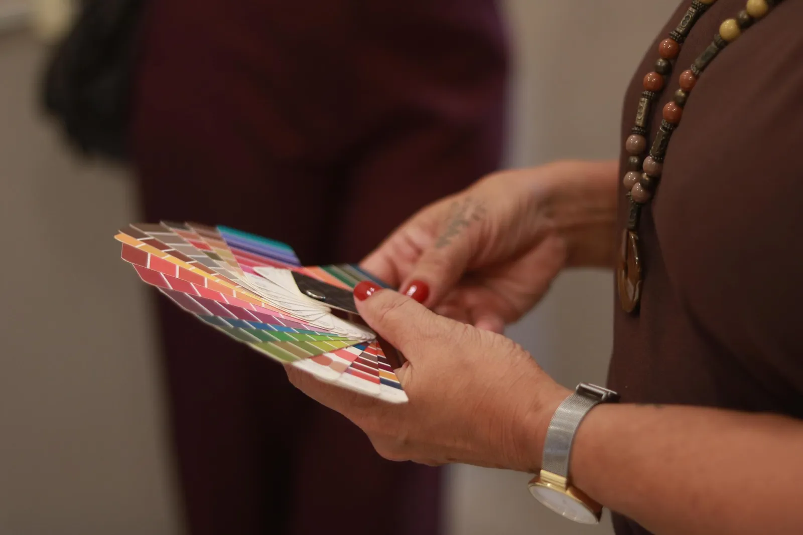 A close-up of hands holding an open fan of colour analysis swatches spanning every shade from warm reds and pinks to cool blues and greens, held by a woman wearing a beaded brown necklace.