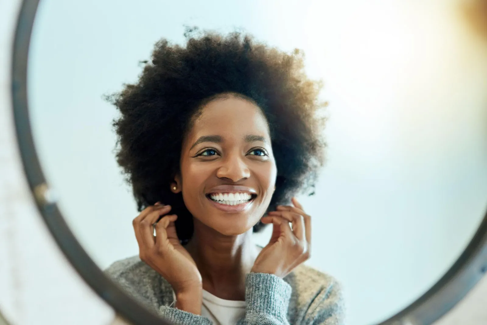 A smiling Black woman with a natural afro reflected in a round mirror, wearing a soft grey cardigan, her face bright and clear in warm light.