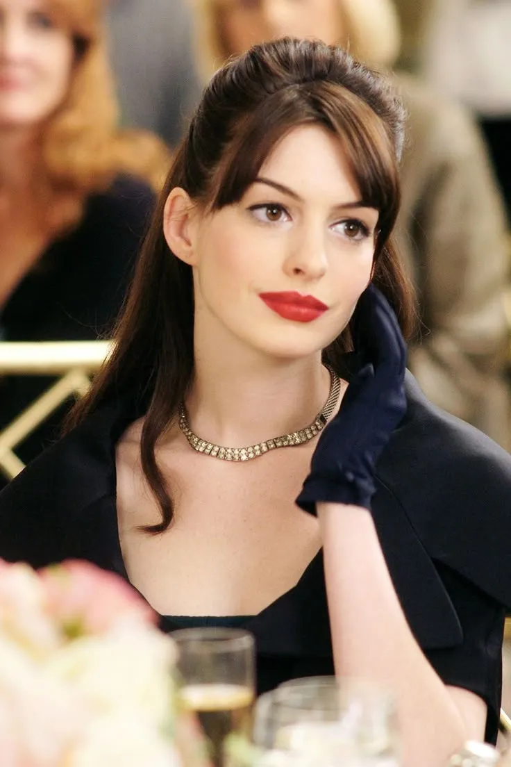 Anne Hathaway in a dark navy jacket with a bold red lip and diamond necklace, seated at an elegant table, her dark hair swept up.