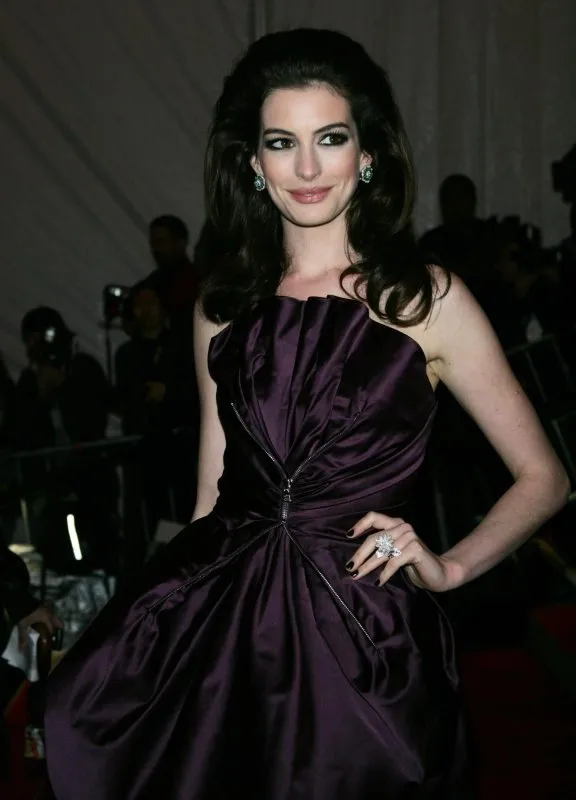 Anne Hathaway in a deep purple aubergine strapless structured gown at a red carpet event, her dark hair voluminous and swept back.