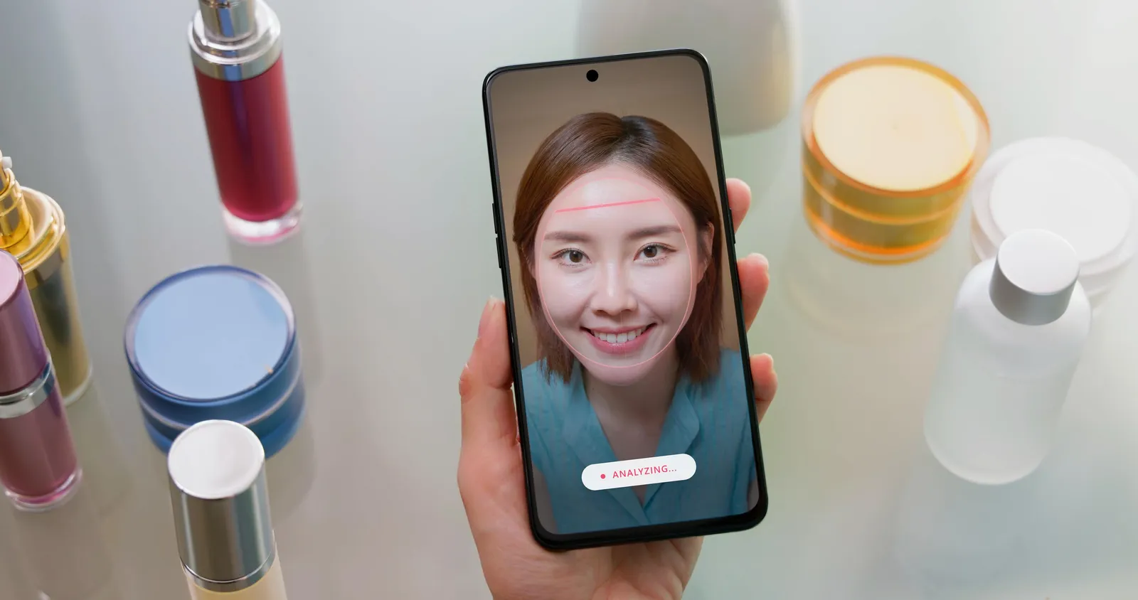 A smartphone held in hand displaying a woman's face being scanned by an AI app, with a red scan line across her forehead and an 'Analyzing...' indicator on screen, surrounded by cosmetic products.