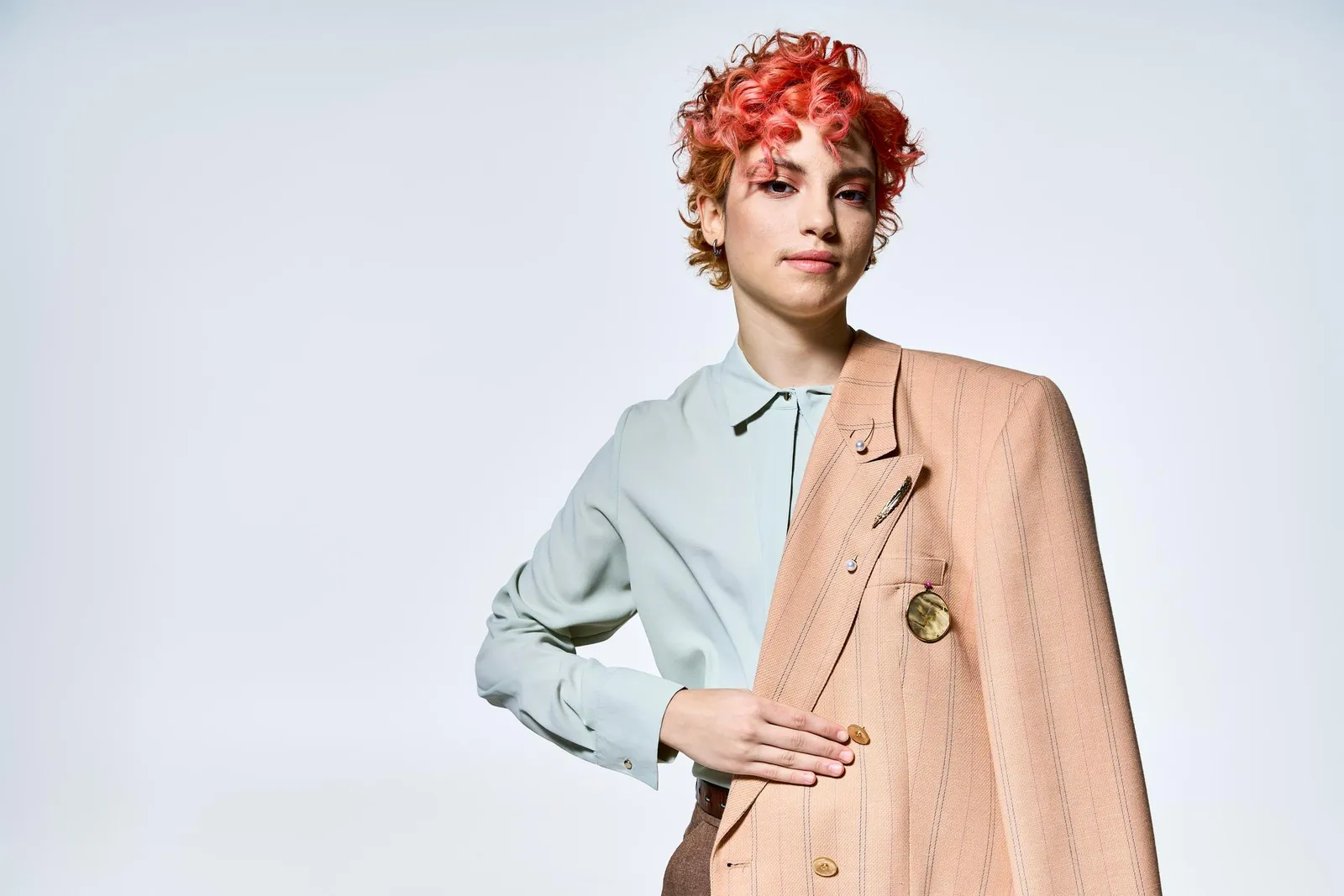 A woman with short curly red hair wearing a pale mint blouse and a peach-toned blazer, photographed against a plain light background.