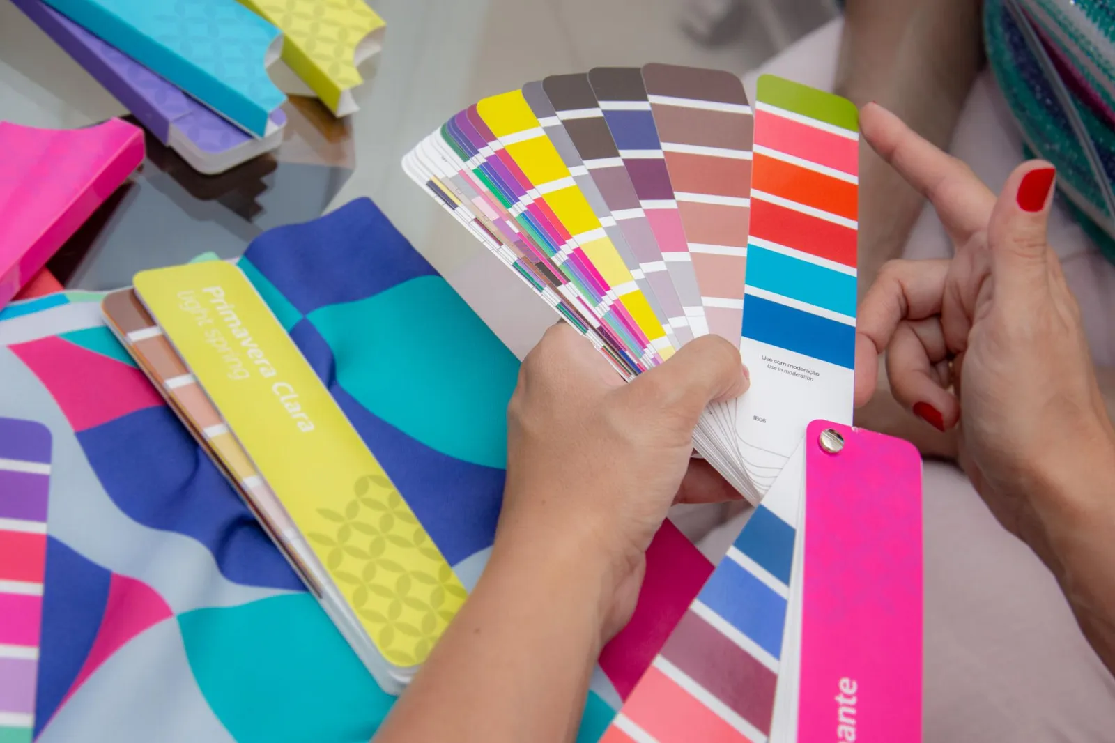Two hands holding a fanned spread of professional colour swatch cards in vivid hues, with additional colour booklets and seasonal palette guides spread across the table.