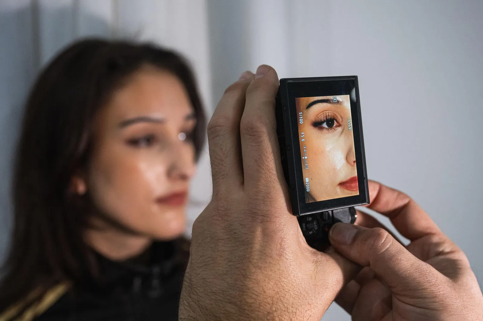 A close-up of a camera LCD screen showing a magnified view of a woman's eye and skin, with the woman herself softly blurred in the background.