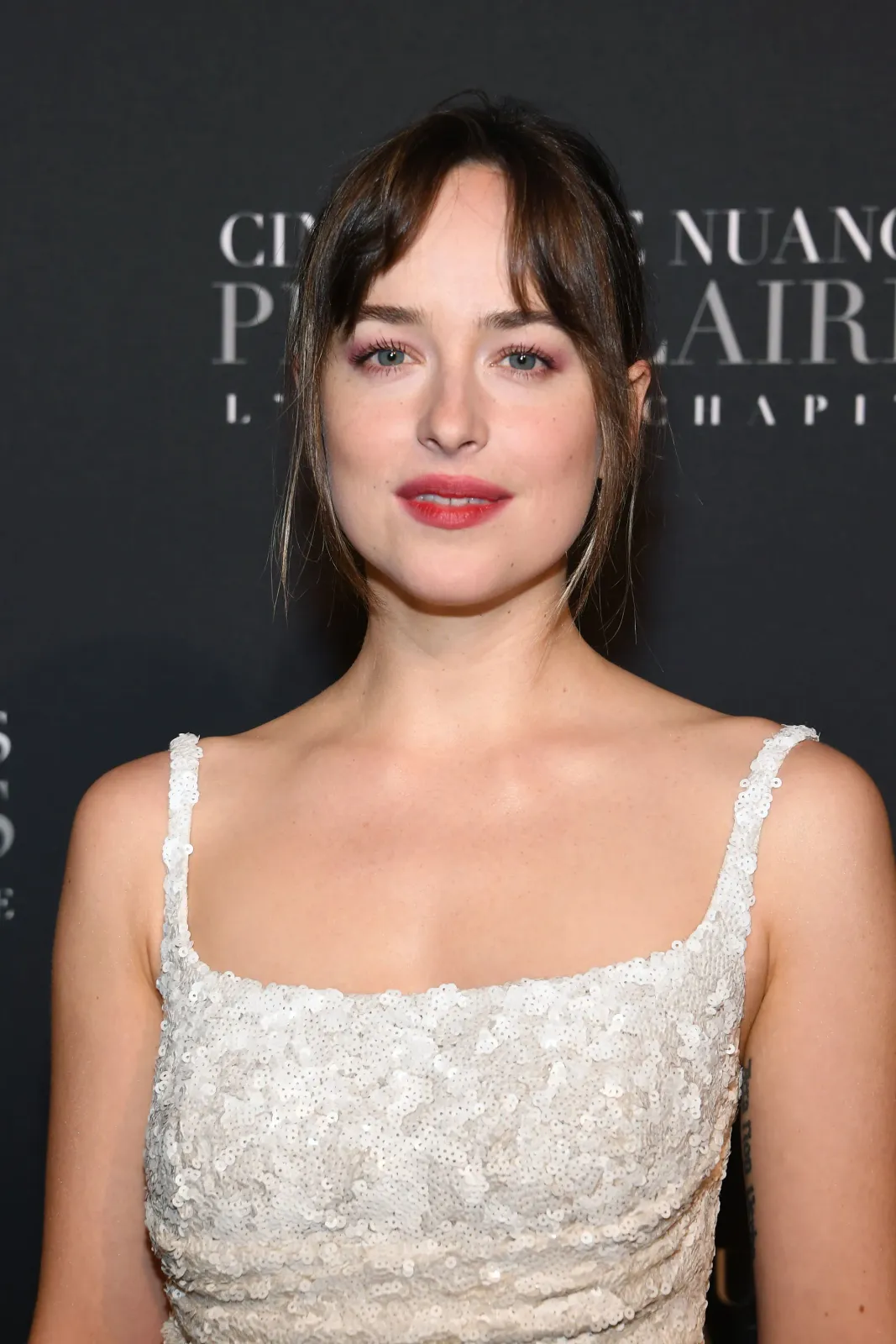 Close-up of Dakota Johnson in a white sequined dress at a premiere, soft pink makeup, dark fringe, against a dark background.