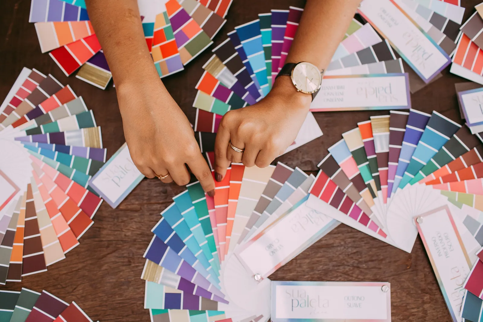 Overhead view of two hands with warm skin tones leafing through dozens of fanned colour swatch booklets spread across a dark wood table.