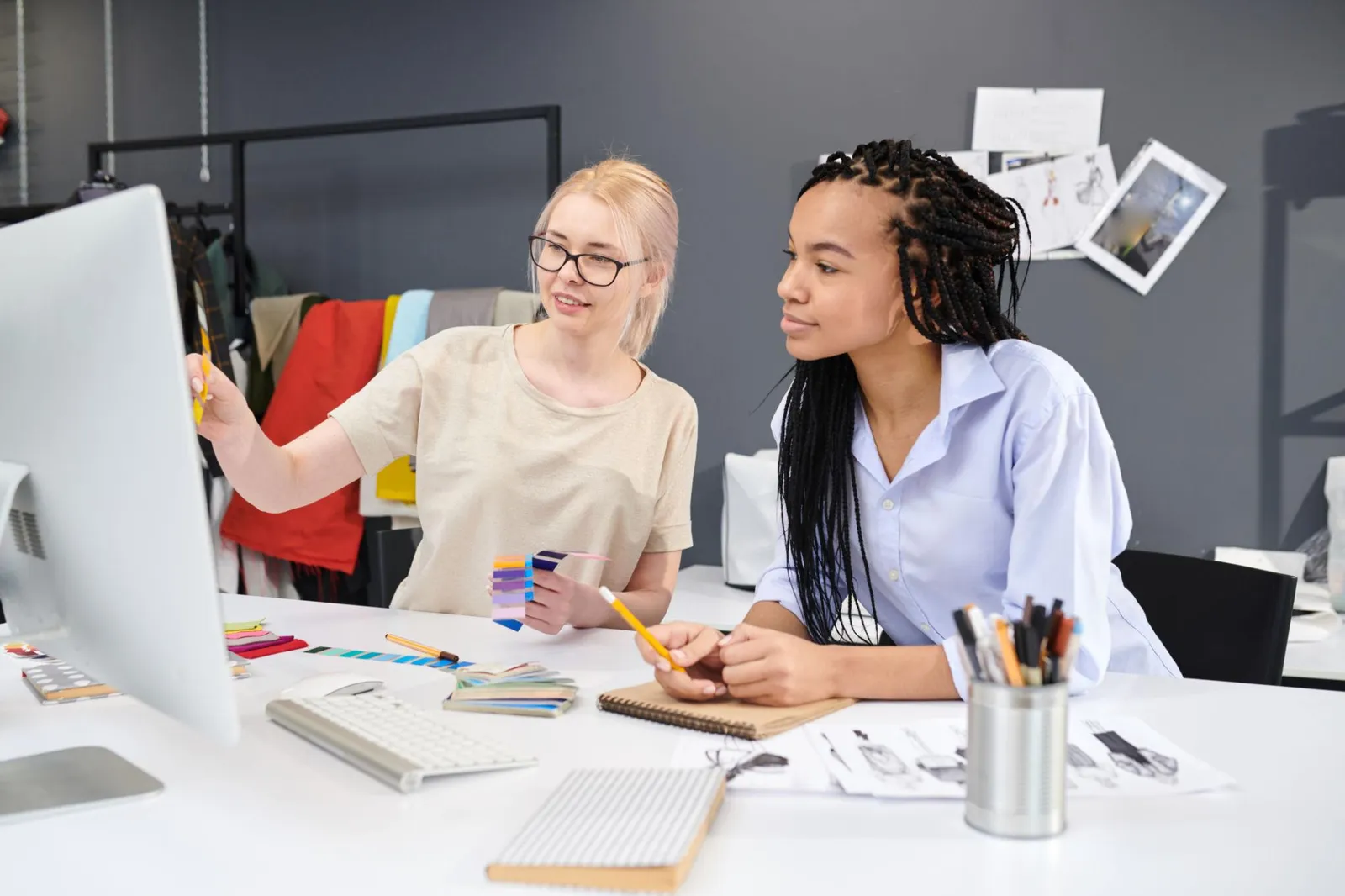 Two women in a fashion design studio reviewing colour swatches together at a desk in front of a monitor, with garments and mood boards visible in the background.
