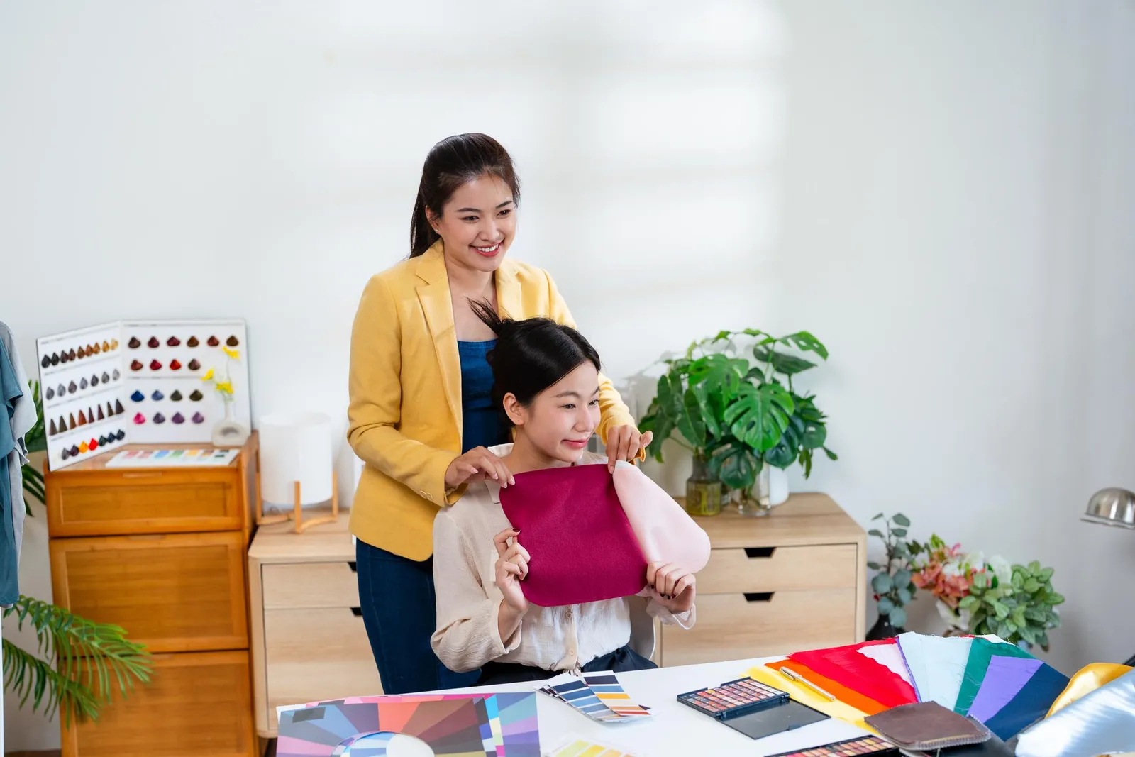 A colour consultant in a yellow blazer draping magenta and blush pink fabric swatches against a seated client's neckline, with colour palettes and swatches spread across the table in front of them.