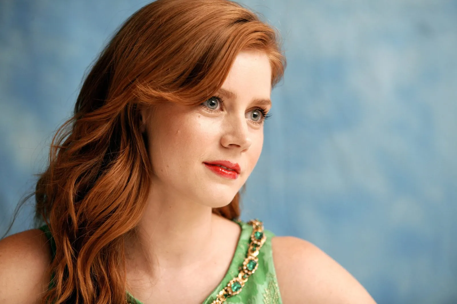 A close-up portrait of Amy Adams with warm copper-red hair worn loose, clear blue-green eyes, peachy skin and a coral lip, wearing a light green embellished top against a blue textured background.