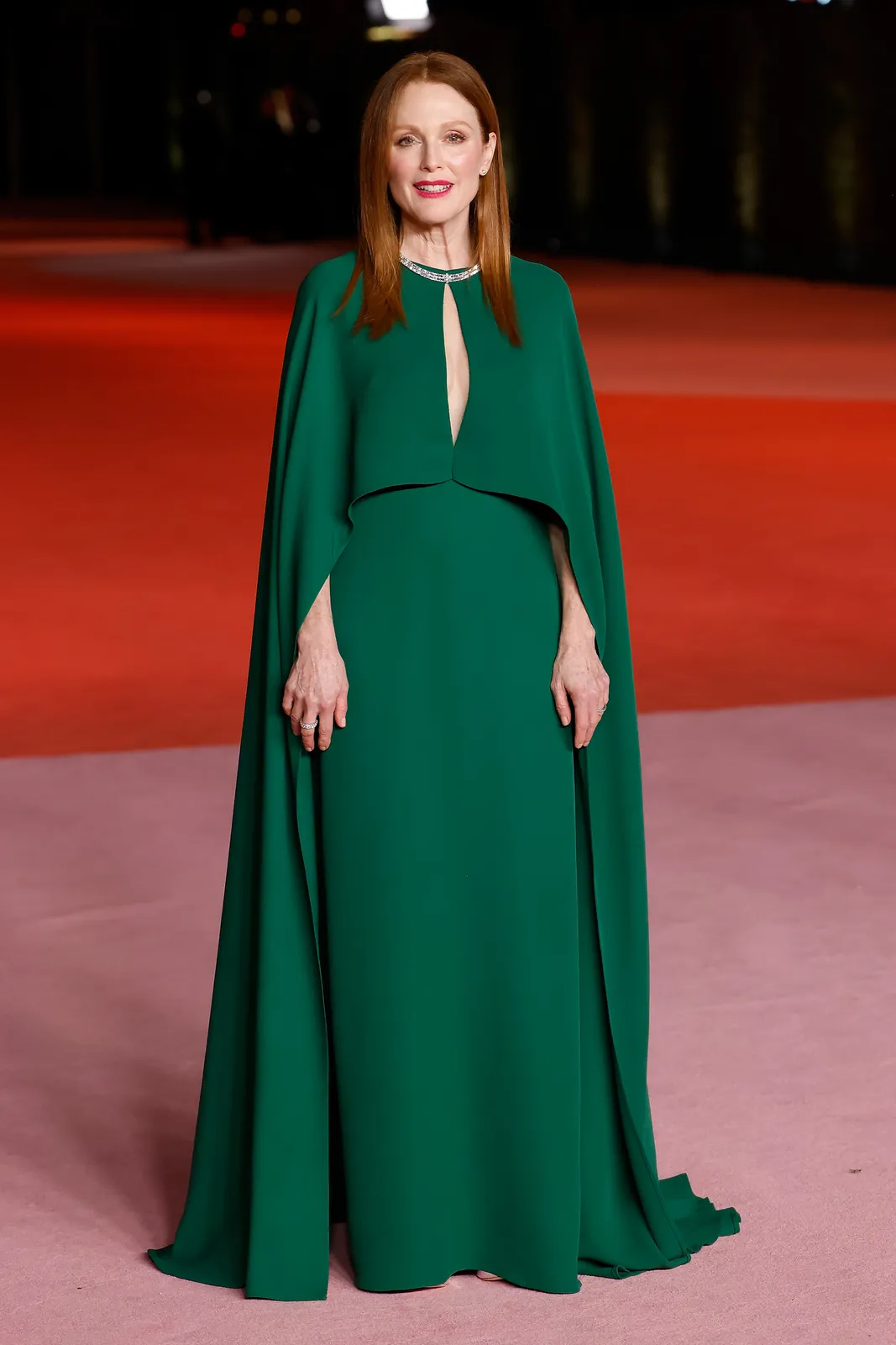 Julianne Moore standing on a carpet in a full-length forest green cape gown with a keyhole neckline, her copper-red hair worn straight, the warm green and her warm colouring perfectly aligned.