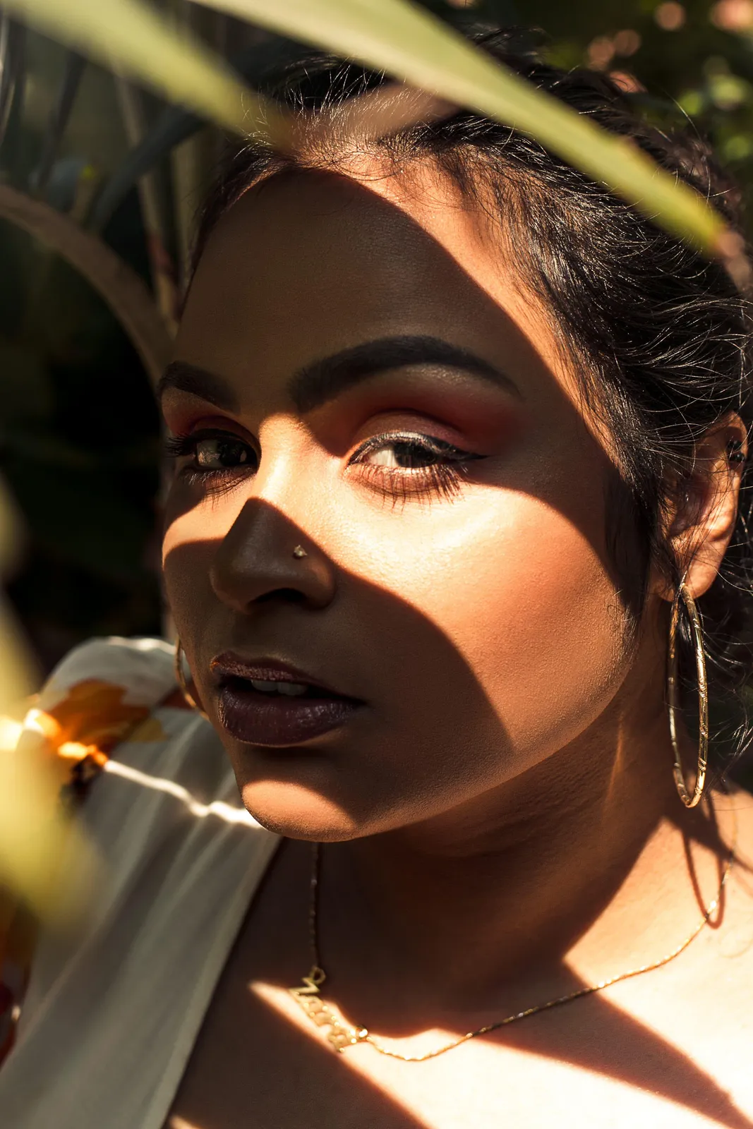 Close-up portrait of a South Asian woman with warm-toned skin, her face lit by dappled sunlight filtering through foliage, gold hoop earrings catching the light.