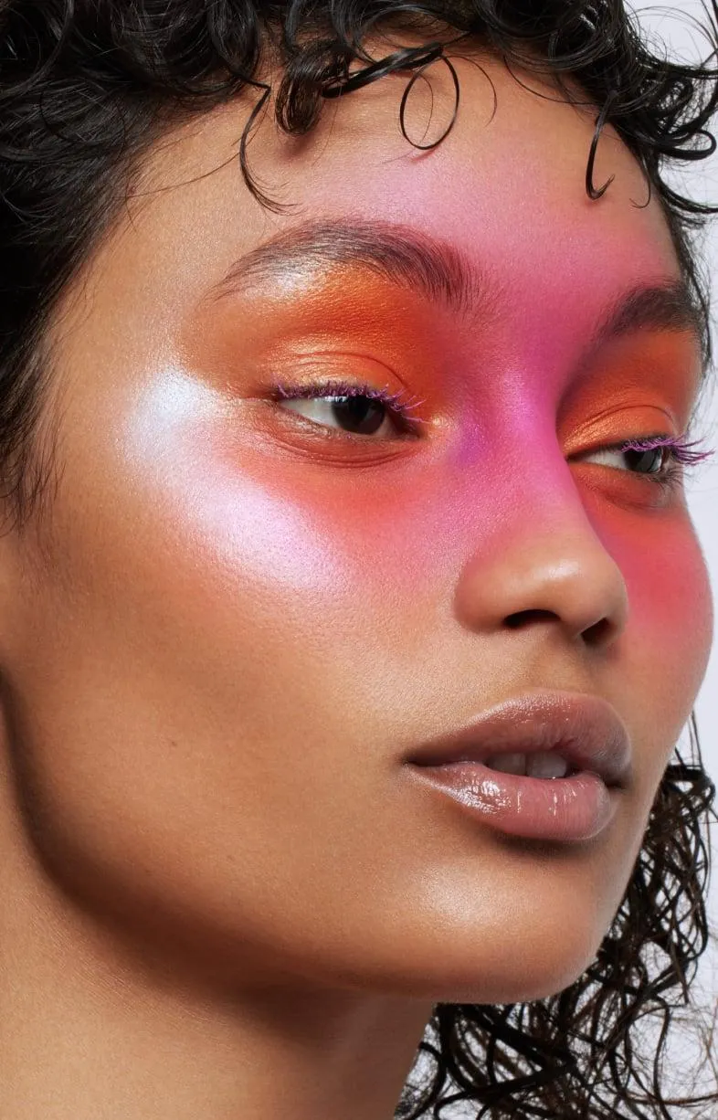 Editorial close-up of a woman with curly dark hair wearing bold warm-toned makeup: vivid orange and pink eyeshadow swept across both eyes, skin glowing with saturation and life.
