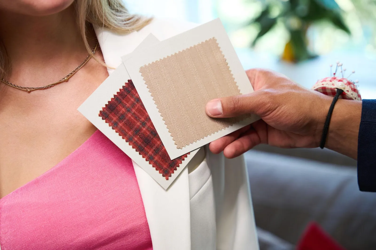 A consultant holds fabric swatches in tartan and beige against the chest of a blonde woman wearing a pink top and white blazer, comparing how each colour reads against her skin.