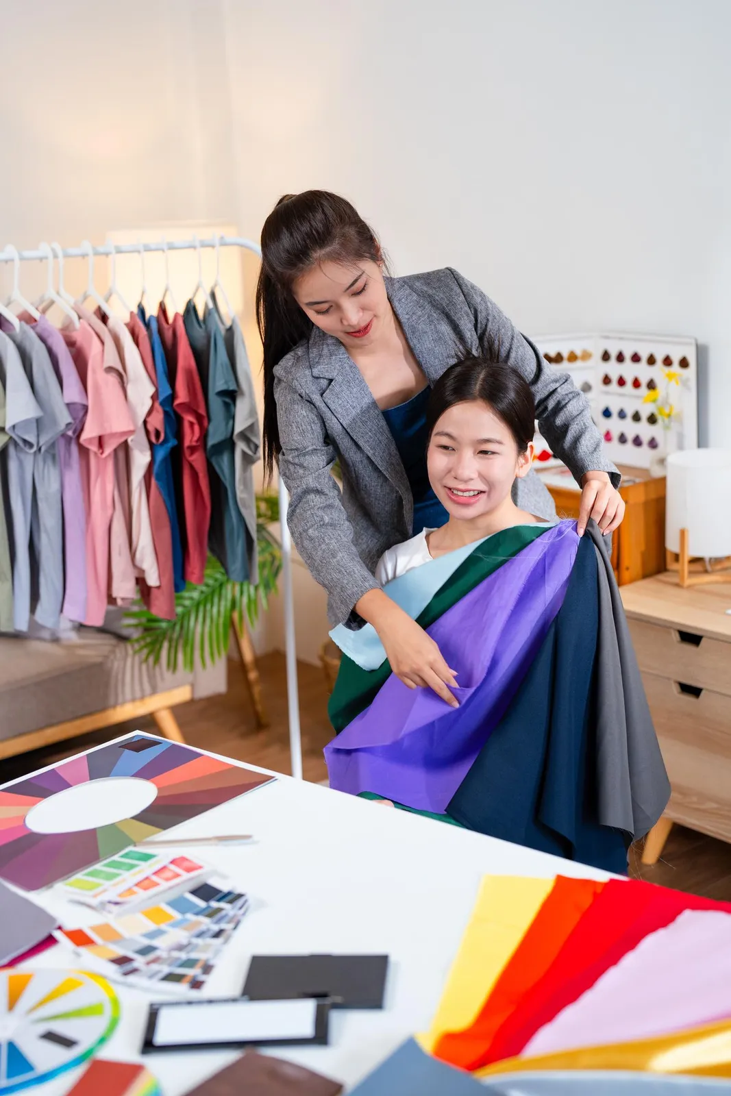 A colour consultant draping purple, teal, and charcoal fabric swatches across the shoulders of a seated client, with a colour wheel, palette cards, and fabric samples spread on the table in front of them.