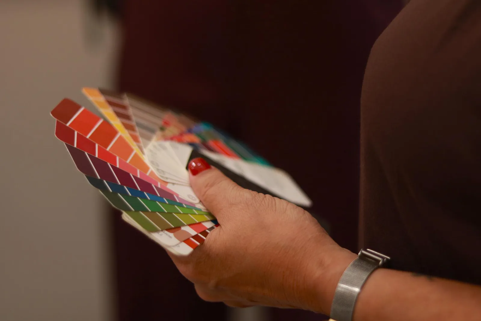 A close-up of a hand holding several fanned colour swatch booklets spanning a full spectrum: reds, oranges, pinks, purples, blues, greens, and yellows arranged in graduated rows.