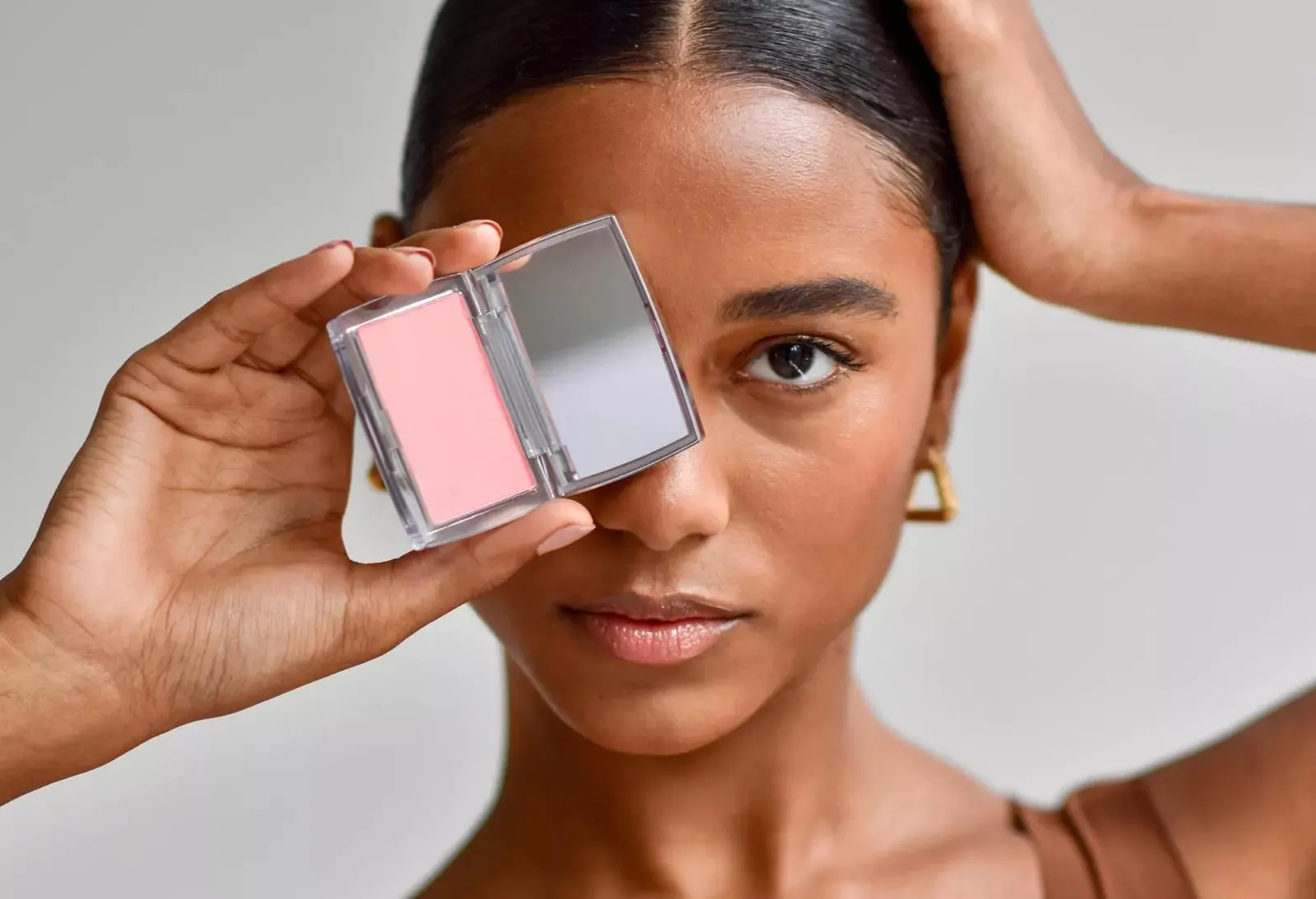 A woman with deep warm-toned skin holds an open compact containing a cool pink blush panel beside its mirror, one eye partially obscured, illustrating how colour choices interact with skin tone.