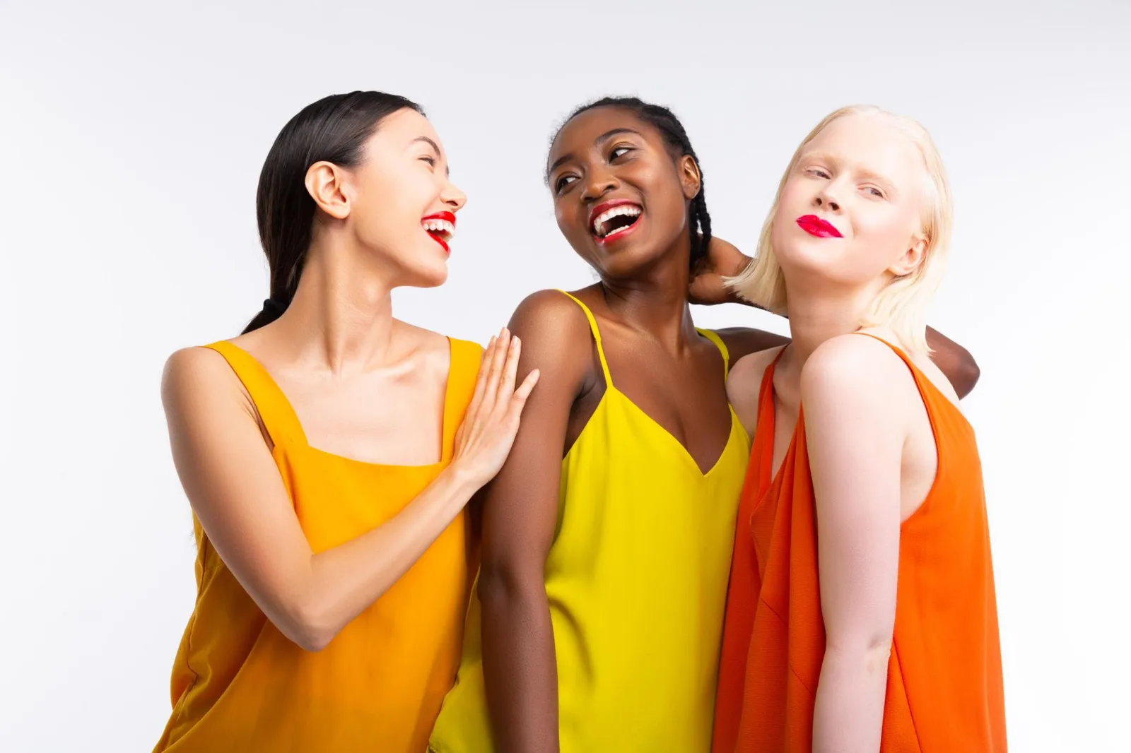 Three women with strikingly different colouring, from warm-toned with dark hair to deep-toned to very fair and blonde, all wearing warm yellow and orange tones and laughing together against a white background.