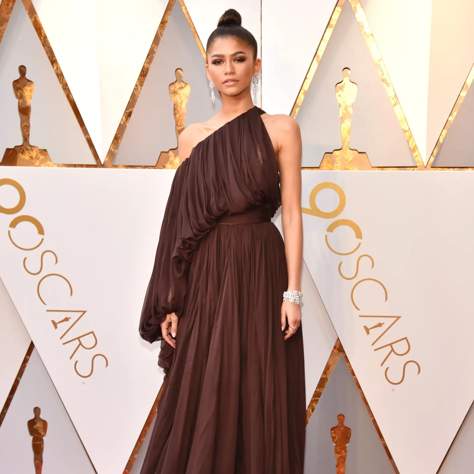 Zendaya at the Oscars in a dramatic deep chocolate-brown one-shoulder draped chiffon gown, hair in a high bun, diamond cuff bracelet, full-length against the gold and white Academy Awards backdrop.