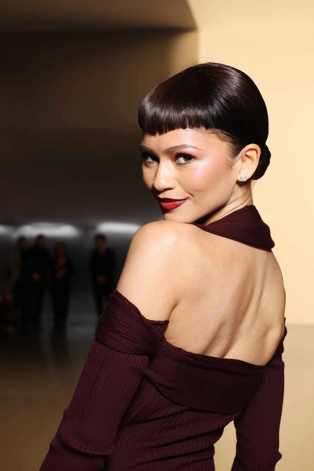 Zendaya looking over her shoulder in a deep aubergine-burgundy off-shoulder ribbed top, wearing a bold red lip and a sleek bowl-cut updo, photographed against a warm neutral background.